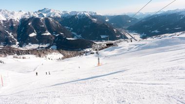 Blick auf eine Skipiste mit einigen Skifahrern, einem Sessellift und den verschneiten Bergen im Hintergrund.