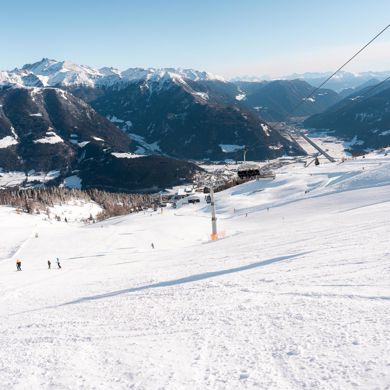 Blick auf eine Skipiste mit einigen Skifahrern, einem Sessellift und den verschneiten Bergen im Hintergrund.