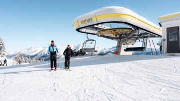 Two skiers in front of a chairlift station in snowy mountains under a bright blue sky.