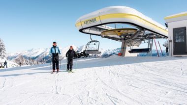 Two skiers in front of a chairlift station in snowy mountains under a bright blue sky.