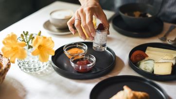 Hand greift nach Chia-Pudding mit Himbeere beim Frühstück, neben Marmelade, Käse und Croissant.