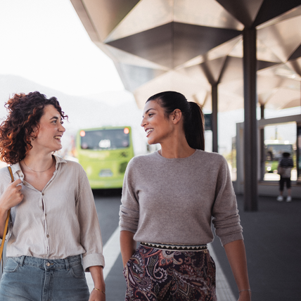 Zwei Frauen unterhalten sich lachend an einer modernen Bushaltestelle mit Blick auf einen grünen Bus im Hintergrund.