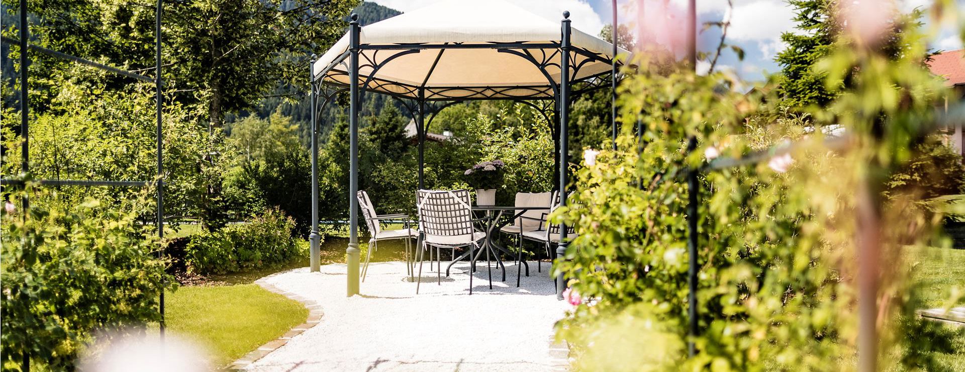Pavillon mit Tisch und Stühlen in einem blühenden Garten bei Sonnenschein, umgeben von Rosen und Grünpflanzen.