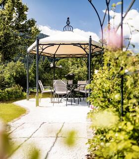 Gazebo con tavolo e sedie in un giardino fiorito sotto il sole, circondato da rose e piante verdi.