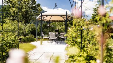 Gazebo con tavolo e sedie in un giardino fiorito sotto il sole, circondato da rose e piante verdi.