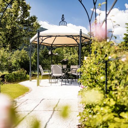 Pavillon mit Tisch und Stühlen in einem blühenden Garten bei Sonnenschein, umgeben von Rosen und Grünpflanzen.