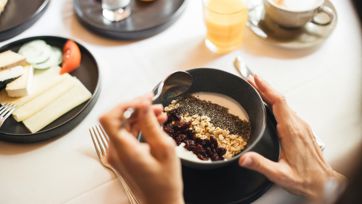 Gesundes Frühstück mit Joghurt, Müsli und Saft auf gedecktem Tisch, Hände mit Löffel sichtbar.