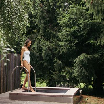 Woman in white swimsuit steps into plunge pool surrounded by lush forest scenery.